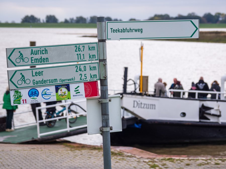 Fahrradwegweiser am Fähranleger Petkum und Fähre Ditzum im Hintergrund.